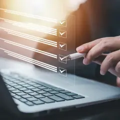 Dłoń z długopisem zaznacza pozycje na liście kontrolnej wyświetlanej nad klawiaturą laptopa.