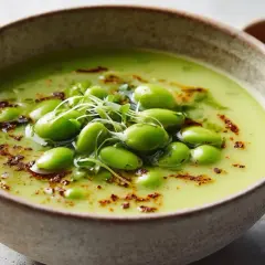 A vibrant green soup, possibly a creamy pea or edamame soup, is garnished with fresh japanese mame beans and microgreens.