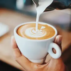 En hand håller en vit kopp med en vacker kaffe latte, där mjölken skapar ett hjärta i skummet.