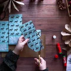 Ręce tną papier z reniferami, tworząc ozdoby choinkowe z papieru. Obok kleje, szyszki i choinka.