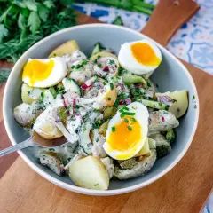Schüssel mit cremigem Kartoffelsalat mit Gurken, Zwiebeln und gekochten Eiern, garniert mit frischen Kräutern.