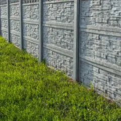 Szary, betonowy płot z fakturą kamienia, obok soczystej, zielonej trawy. Idealny do malowania i odświeżenia.