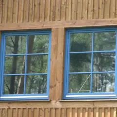Dwa niebieskie okna ze szprosami w drewnianej elewacji. Odbijają się w nich drzewa.