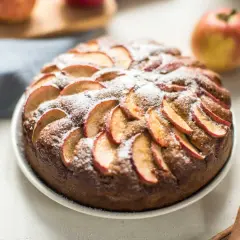 Un délicieux moelleux aux pommes, saupoudré de sucre glace, avec des pommes fraîches et des bâtons de cannelle en arrière-plan.