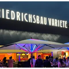 Leuchtende Buchstaben über dem Eingang des Varieté Friedrichsbau. Menschen versammeln sich unter beleuchteten Sonnenschirmen.