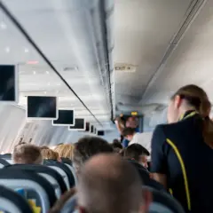 Widok z kabiny samolotu. Stewardka w granatowym mundurku z żółtym pasem obsługuje pasażerów.
