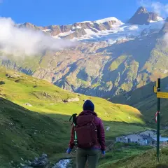 A hiker pauses on a **Tour Mont Blanc hike**, gazing at a majestic snow-capped mountain range. A signpost points the way to the next stage of this epic journey.