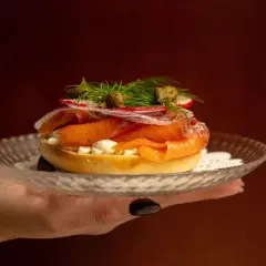 En hand håller upp en bagel med rökt lax, färskost, rödlök, rädisor och kapris. Perfekt för en lyxig bank hotel middag.