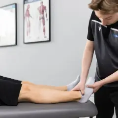 Fizjoterapeuta w sporcie delikatnie bada stopę zawodnika leżącego na kozetce.