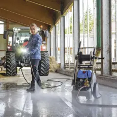Frau reinigt Stallboden mit einem Industrie Hochdruckreiniger. Kühe und Traktor im Hintergrund.