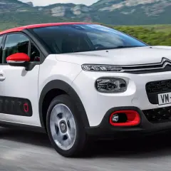 Biały Citroen C3 2025 z czerwonym dachem i lusterkami jedzie po drodze, otoczony zielonymi wzgórzami.