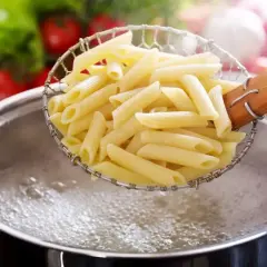 Penne makaron al dente w cedzaku nad garnkiem bulgoczącej wody. W tle pomidory i sałata.