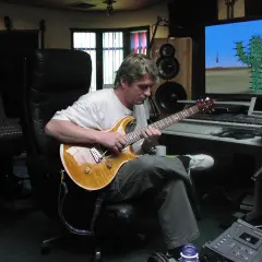 Mike Oldfield w studiu, gra na gitarze. Na ekranie komputerowym kaktus na pustyni, jak w jego utworach.