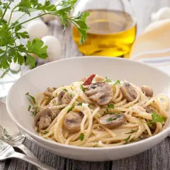 Talerz spaghetti z pieczarkami, posypany parmezanem i natką pietruszki. Obok sztućce, oliwa i zioła.