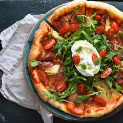 Pyszna pizza z burrata, rukolą, pomidorkami koktajlowymi i szynką parmeńską, podana na niebieskim talerzu.