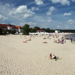 Plaża w Sopocie z ludźmi opalającymi się i spacerującymi, w tle budynki i morze.