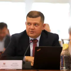 Jacek Wójcicki, siedząc przy stole, przemawia. Jego wzrost jest przeciętny, a na stole leży laptop i woda z cytryną.