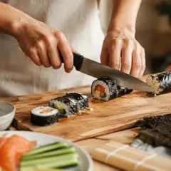 Ręce kroją rolkę sushi na desce. Widać składniki: ryż, łososia, ogórka. Pyszne domowe sushi w przygotowaniu.