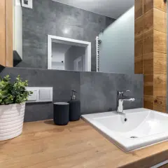 Drewniany blat w łazience z umywalką, baterią, dozownikiem mydła i rośliną w doniczce.