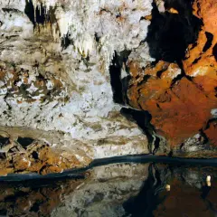 Interior de una cueva con estalactitas y formaciones rocosas rojizas, reflejándose en un espejo de agua. El **cerro del hierro** se revela en su esplendor subterráneo.