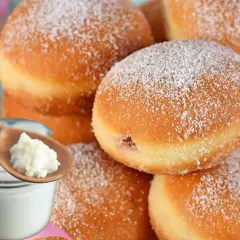 Puszyste pączki z lukrem, posypane cukrem pudrem, obok słoiczka z twarożkiem i drewnianej łyżeczki.