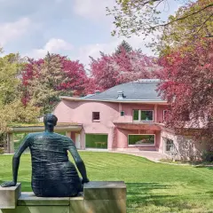 Bronze-Figur blickt auf rosafarbenes Haus im Skulpturenpark Waldfrieden, umgeben von leuchtend rotem und grünem Laub.