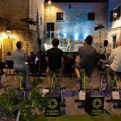 Spettatori pedalano su cyclette per alimentare un'esibizione di teatro a pedali in una piazza illuminata.