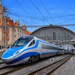 Nowoczesne oznaczenie Pendolino na tle zabytkowego dworca w Pradze. Pociąg w kolorach niebieskim i srebrnym stoi na torach.