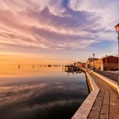 Zachód słońca nad laguną w Wenecji. Spokojna woda odbija różowo-pomarańczowe niebo. Wzdłuż kanału ciągnie się kamienna promenada z latarniami.
