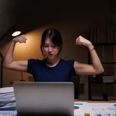 Młoda kobieta w nocy przy biurku pokazuje bicepsy, jakby cieszyła się z dodatku za pracę w nocy.