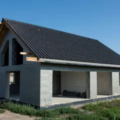 Dom w stanie surowym otwartym, z dachem pokrytym czarną dachówką. Widoczne otwory okienne i drzwiowe, betonowe ściany.