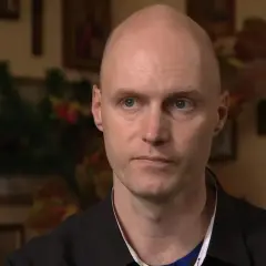 Krzysztof Igor Krawczyk, łysy mężczyzna z niebieskimi oczami, w czarnej koszuli, na tle rodzinnych zdjęć.
