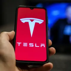Dłoń trzyma telefon z logo Tesla. W tle widać niewyraźną postać na niebieskim tle, być może Elon Musk.
