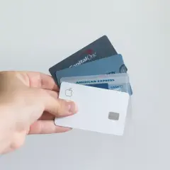 Dłoń trzyma kilka kart płatniczych, w tym Apple Card, American Express i Capital One. Standardowe wymiary karty bankomatowej są widoczne.