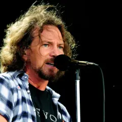Eddie Vedder, wokalista Pearl Jam, śpiewa do mikrofonu na koncercie. Jego rozwichrzone włosy i charakterystyczna koszula dodają mu rockowego stylu.