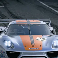 Srebrny Porsche 918 Spyder z pomarańczowymi pasami i numerem 22 na masce pędzi po torze wyścigowym.