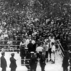 Boxing ring with fighters and officials, surrounded by a massive, cheering crowd in vintage attire.