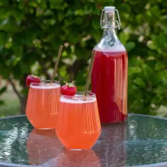 Två glas med rosa rabarbersaft, garnerade med jordgubbar och sugrör, står på ett glasbord. En flaska med saft i bakgrunden. Perfekt för ett rabarbersaft recept.