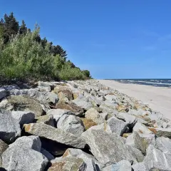 Kamienisty brzeg morza z zielenią i piaszczystą plażą w tle.