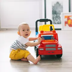 Uśmiechnięte niemowlę w żółtych spodenkach bawi się czerwono-żółtą ciężarówką na drewnianej podłodze.
