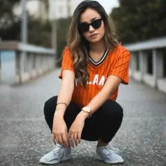 Chica con gafas de sol y camiseta naranja a rayas, pantalones negros y zapatillas deportivas. Un ejemplo de estilos de ropa urbana y moderna.