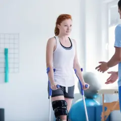 Kobieta na kulach z ortezą na nodze podczas zabiegów rehabilitacyjnych, wspierana przez fizjoterapeutę.