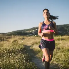 Kobieta w stroju sportowym biegnie po ścieżce wśród traw, w tle wzgórza.