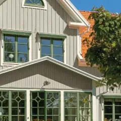 Vitt hus med gråa knutar och orange tak. Gröna fönster och en stor lönn skänker skugga.