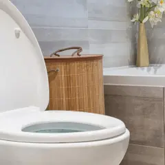Biała deska WC z otwartą klapą, obok bambusowy kosz i wazon z kwiatami. Dowiedz się, jak dobrać deskę WC do swojej łazienki.