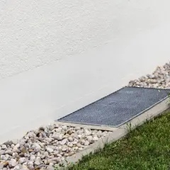 A gravel bed and grate next to a white wall, showing how to level ground for drainage.