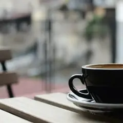 En svart kopp med lungo kaffe står på ett träbord. I bakgrunden syns en bänk och en suddig stadsmiljö.