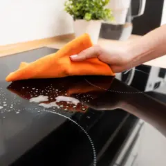 Mano limpiando placa de inducción con paño naranja y espuma. Se puede limpiar la placa de inducción con vitroclen para dejarla reluciente.