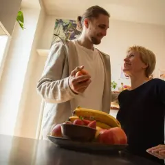 Ein Mann hält einen Apfel, während er mit einer lächelnden Frau spricht. Obst liegt auf einem Tisch. Vielleicht ein Moment der Entlastung für eine Privatperson.
