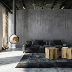 Nowoczesny salon z betonowymi ścianami i podłogą. Centralnym punktem jest wiszący kominek, a obok niego szara kanapa i dwa drewniane stoliki. Surowość formy podkreśla industrialny charakter wnętrza.